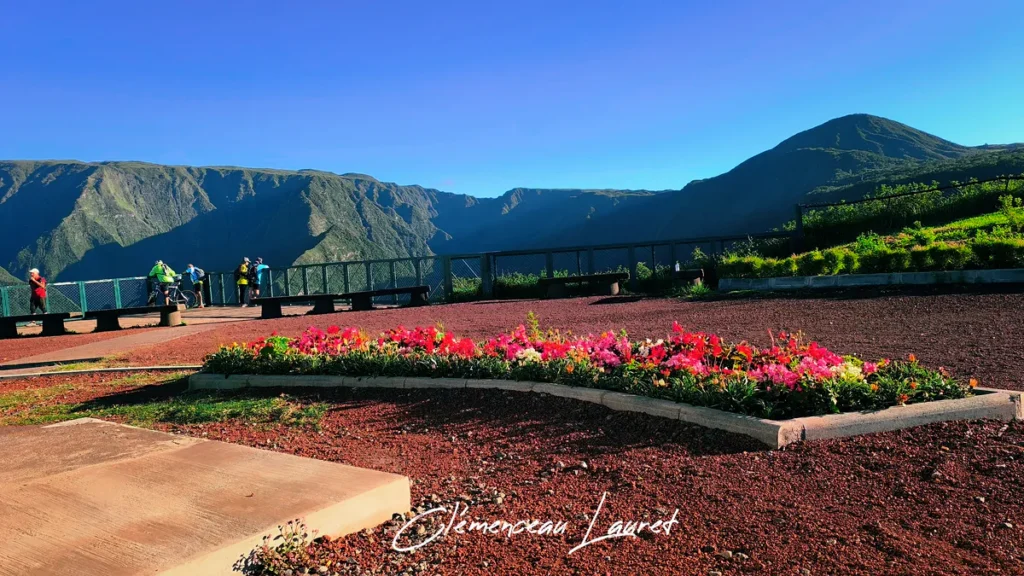 Vue panoramique depuis le Belvédère de Bois Court à la Réunion, montrant des parterres de fleurs, des randonneurs et des montagnes verdoyantes.