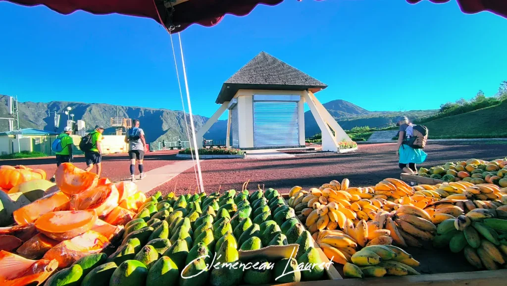 Un marché coloré au Belvédère de Bois Court à la Réunion, avec des avocats, des kakis, des citrouilles et des bananes exposés sous un parasol rouge.
