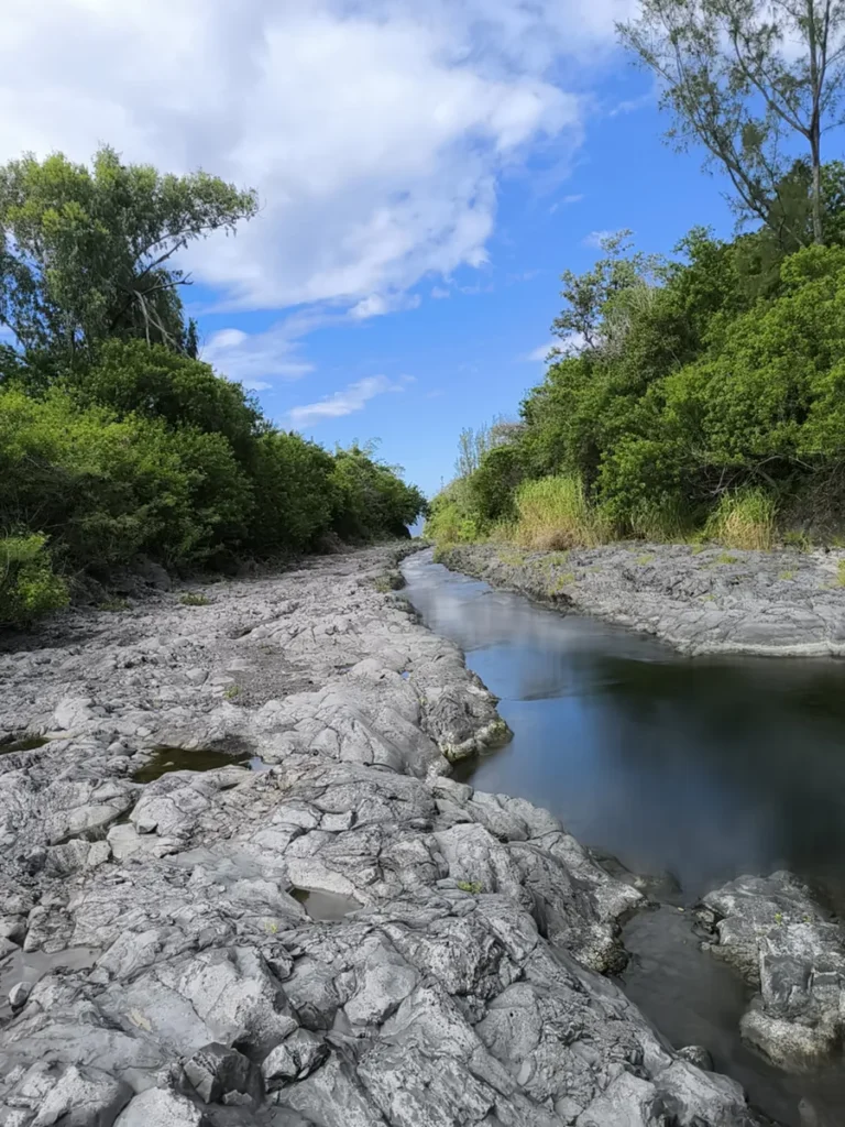 Vue en contre-plongée du lit rocheux en basalte gris traversé par un filet d'eau sombre sous un ciel bleu nuageux à la rivière Langevin.