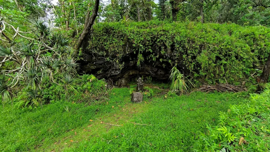 Vue d'ensemble de l'abri sous roche basaltique envahi par la végétation avec la statue de Lou Caf à l'entrée.