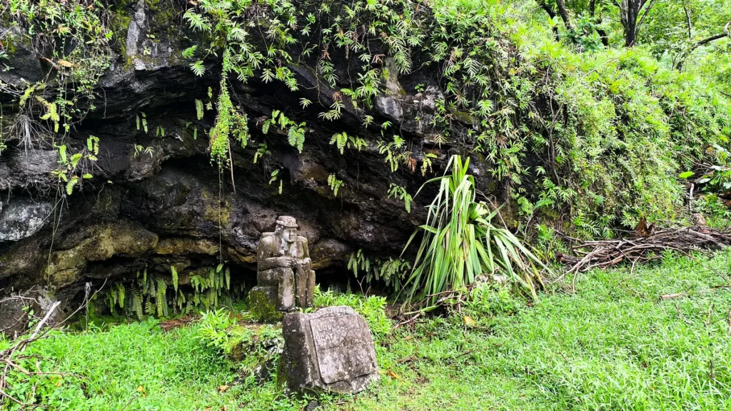Statue en pierre sculptée d'un homme assis devant la cavité rocheuse recouverte de fougères à Sainte-Rose.