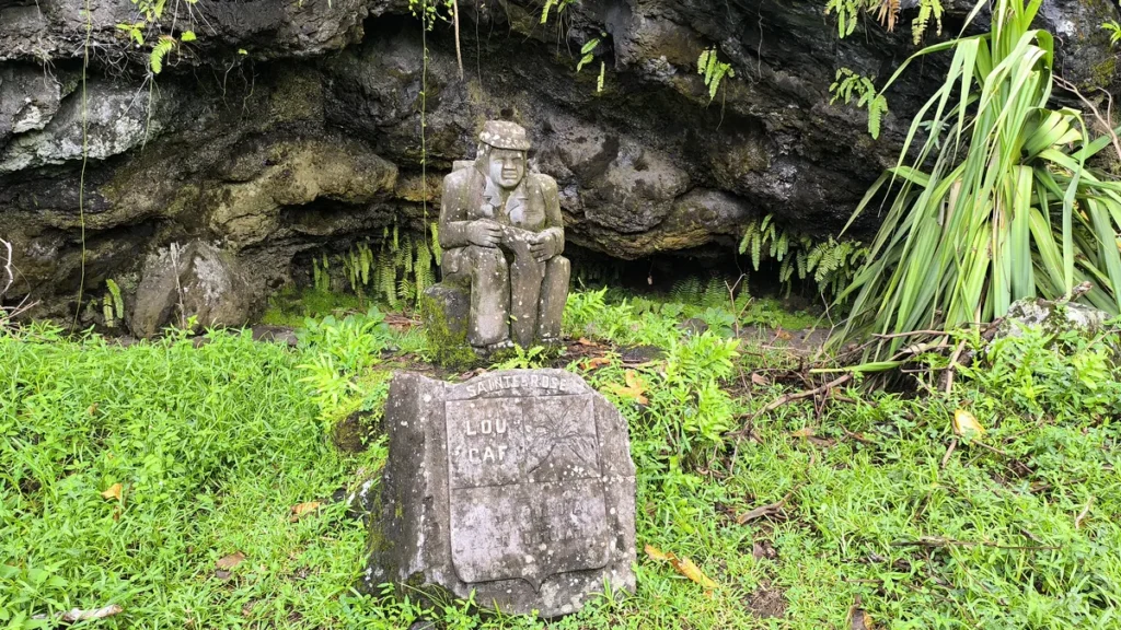 Angle frontal montrant le blason gravé et la statue de Lou Caf assis devant la falaise basaltique.