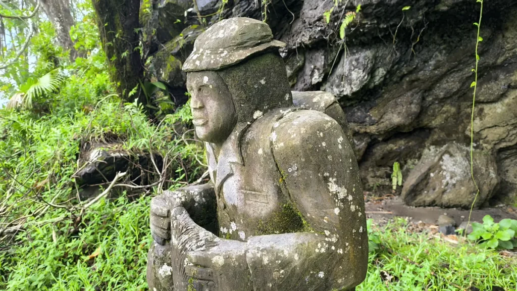 Vue de profil de la sculpture en pierre de Lou Caf portant un chapeau et un sac à dos à Sainte-Rose.