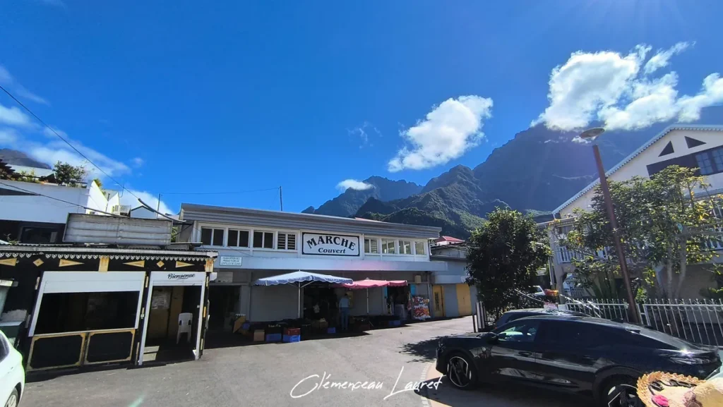 Façade du Marché Couvert de Cilaos avec des parasols et des voitures garées sur la place du village sous un ciel dégagé.