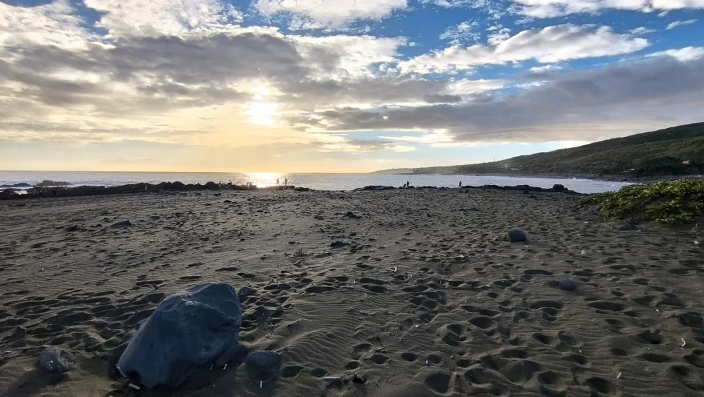 Vue panoramique de la plage de sable noir de l'Étang-Salé couverte de traces de pas, avec de l'eau résiduelle reflétant le soleil couchant.