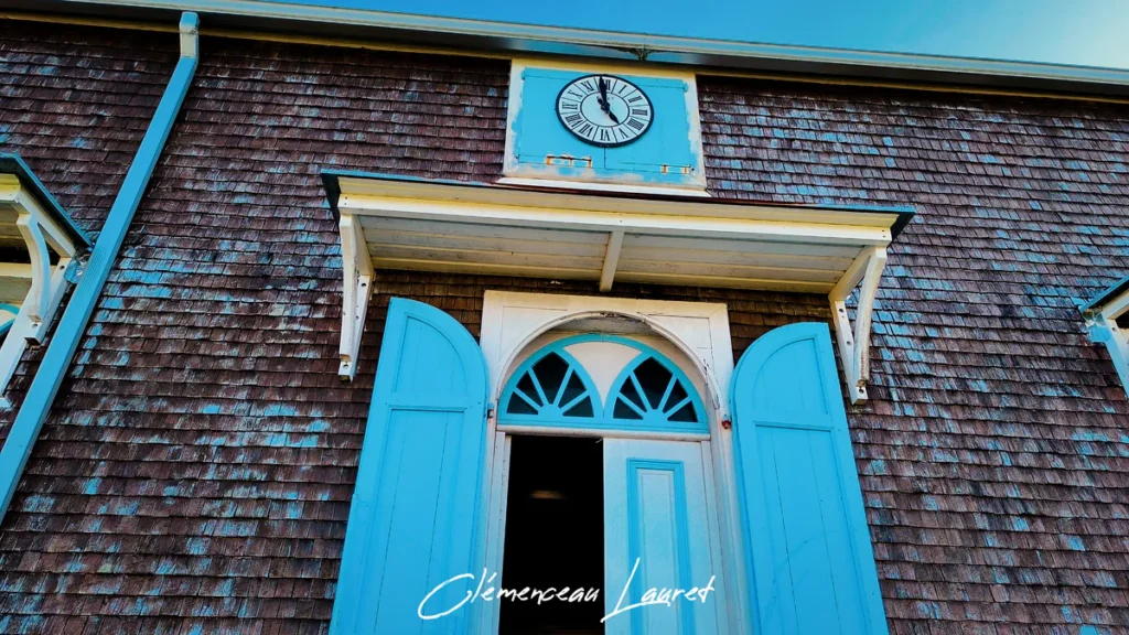 Entrée latérale en bois bleu cyan de l'église des Lianes avec horloge à chiffres romains fixée sur le bardage.