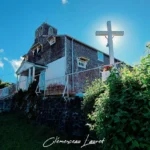 Vue extérieure globale en contre-plongée de l'église des Lianes à Saint-Joseph (La Réunion) avec la grande croix blanche au premier plan, baignée par la lumière du soleil.
