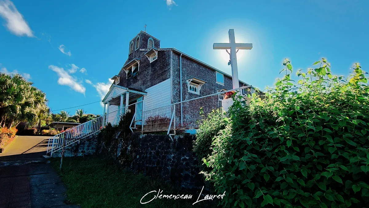 Vue extérieure globale en contre-plongée de l'église des Lianes à Saint-Joseph (La Réunion) avec la grande croix blanche au premier plan, baignée par la lumière du soleil.