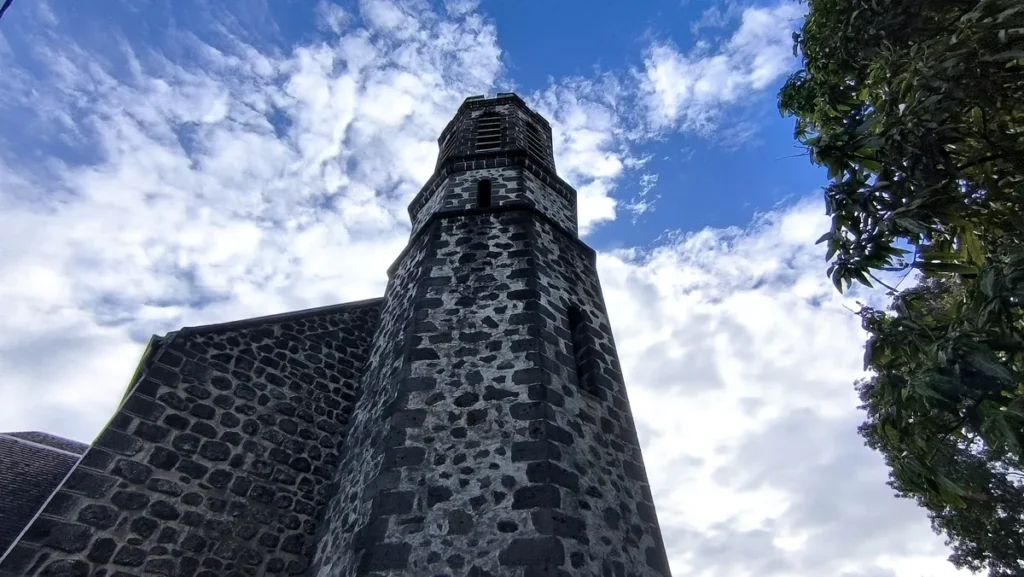 Clocher en pierre de taille sombre de l'église Notre-Dame-de-la-Salette vu en contre-plongée sous un ciel nuageux.