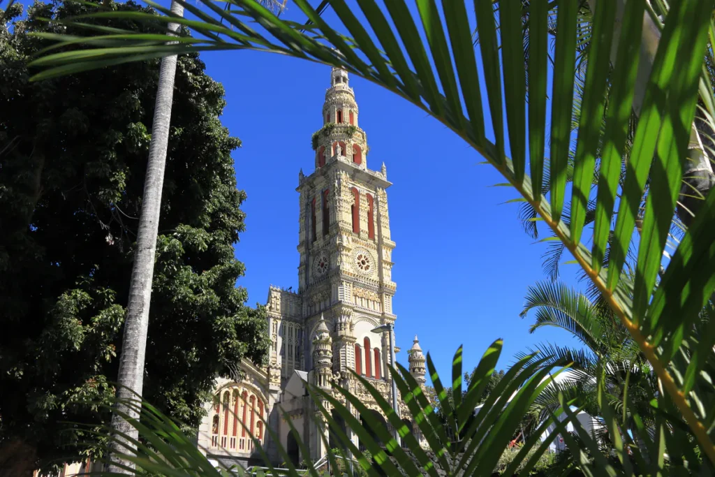 Église Sainte-Anne de La Réunion, avec son clocher orné et ses touches de rouge, vue à travers des palmes de palmier vert sous un ciel bleu vif. Photographie libre de droits.