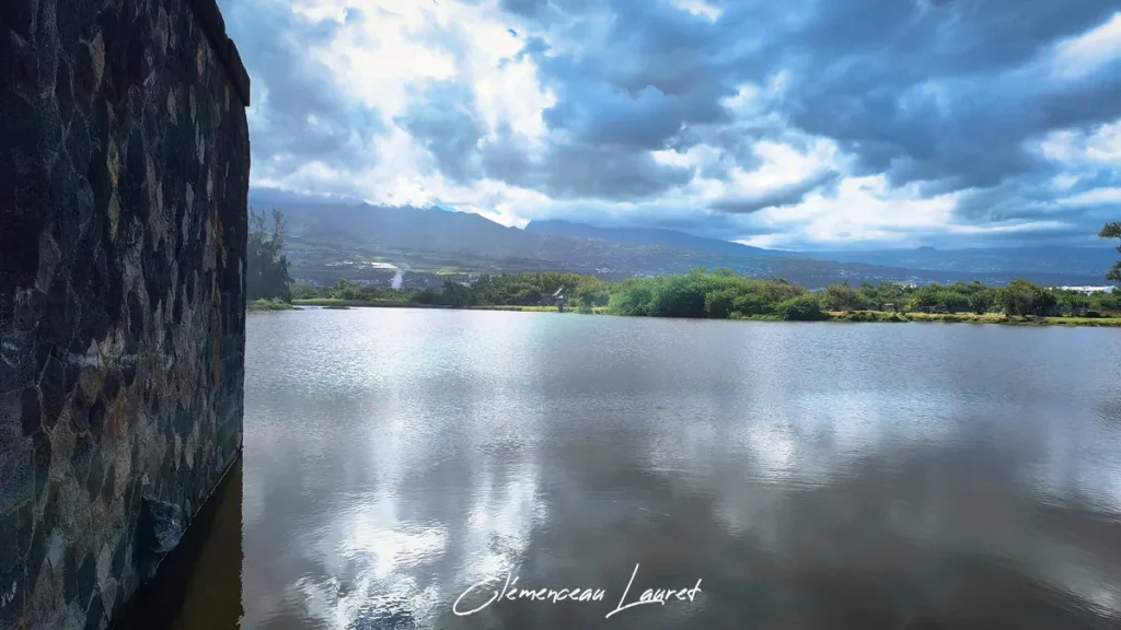 Vue sur le plan d'eau calme de l'Étang du Gol à Saint-Louis avec un mur en pierre de taille au premier plan et des montagnes sous des nuages.