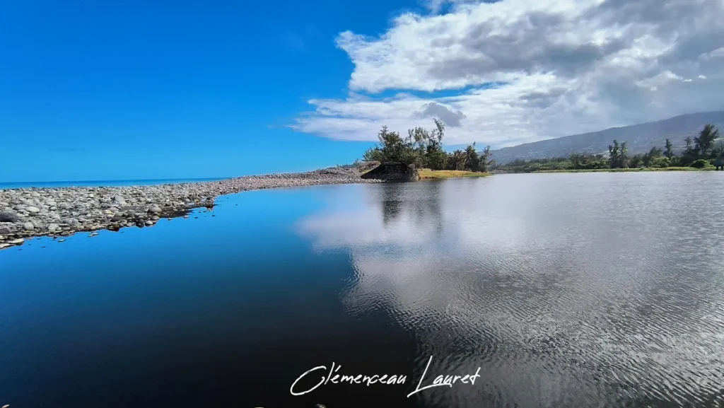 Cordon de galets longeant une eau sombre et calme reflétant le ciel bleu et les nuages à l'Étang du Gol.