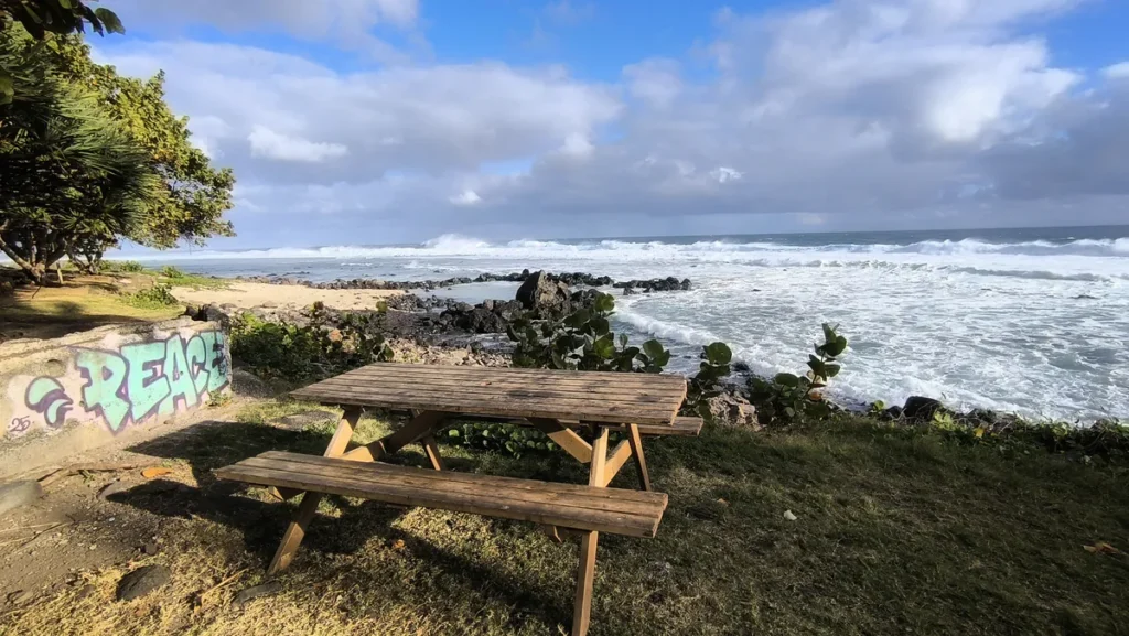 Vue grand-angle d'une table de pique-nique en bois au premier plan, devant un muret tagué "PEACE", surplombant le lagon agité de Grand Bois et ses vagues écumantes sous un ciel nuageux.