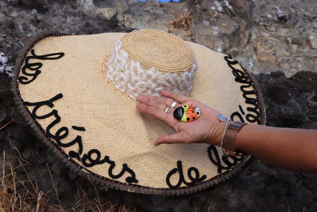 Vue de haut d'un grand chapeau de paille tressé portant l'inscription « Les trésors de la Réunion » avec une main posant le galet coccinelle en son centre.