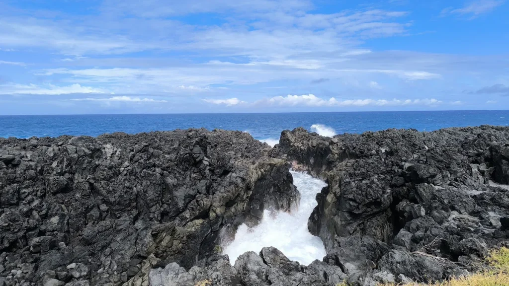 Vague s'engouffrant avec force dans une faille de roche volcanique noire au Gouffre de l'Étang-Salé sous un ciel bleu.