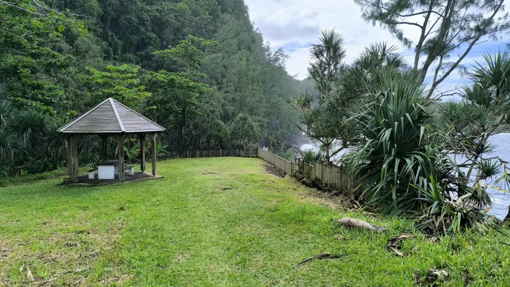 Kiosque en bois sur pelouse verdoyante bordée par des vacoas et la mer à La Source Sainte-Rose.