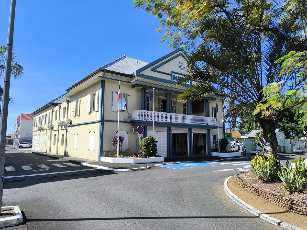 Batiment à deux étages de la mairie de Sainte-Suzanne bordé par une rue goudronnée et des palmiers sous un ciel bleu.