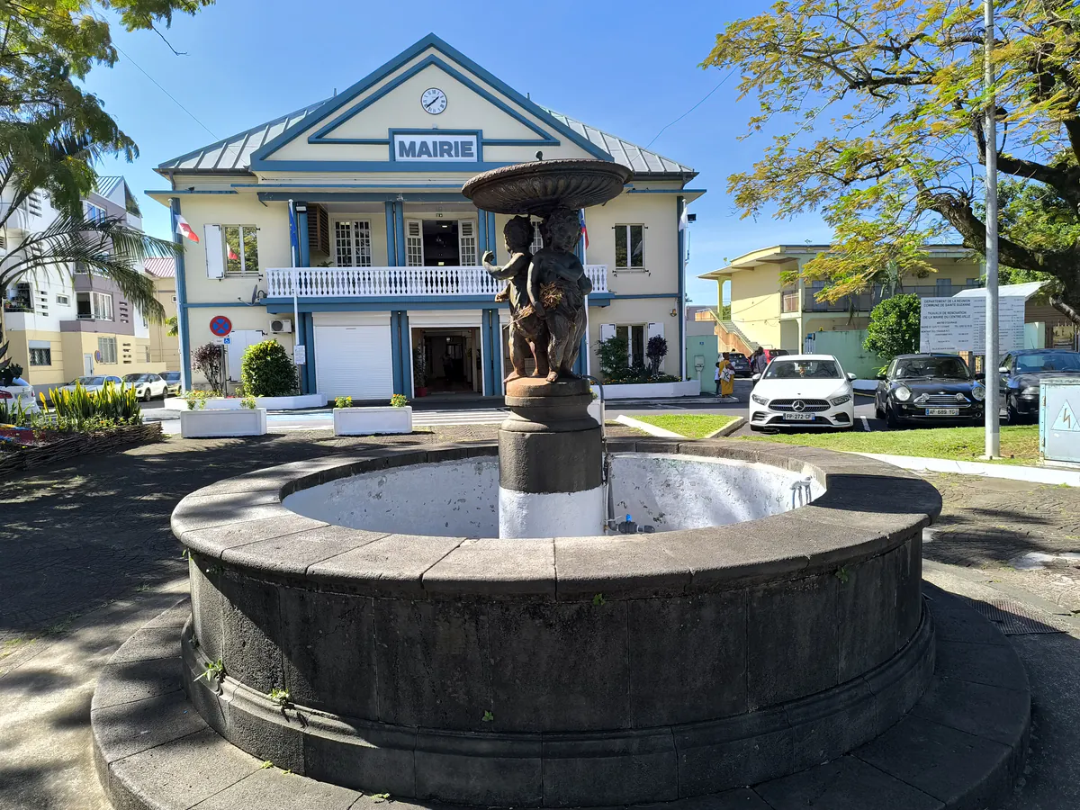 Fontaine en pierre sculptée au premier plan sur une place ombragée avec le bâtiment de la mairie de Sainte-Suzanne en arrière-plan.