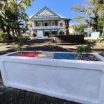 Bac à fleurs rectangulaire blanc garni aux couleurs du drapeau français, avec la fontaine et la mairie de Sainte-Suzanne en arrière-plan.