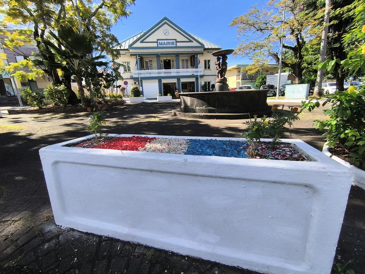 Bac à fleurs rectangulaire blanc garni aux couleurs du drapeau français, avec la fontaine et la mairie de Sainte-Suzanne en arrière-plan.
