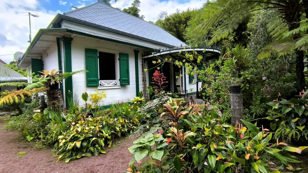 Façade principale de la maison créole avec ses volets verts, son lambrequin et ses massifs de fleurs multicolores.