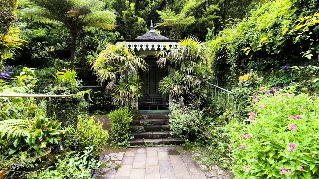 Kiosque créole en bois avec lambrequins ciselés entouré de palmiers et de plantes fleuries.