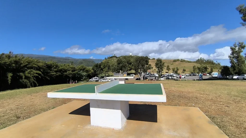 Table de ping-pong en béton installée sur une esplanade en plein air près du parking.