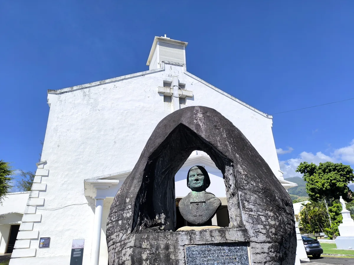 Buste sculpté dans une cavité rocheuse devant la façade blanche de l'église Saint-François-Xavier sous un ciel dégagé.