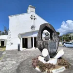 Monument en pierre sculptée sur le parvis bitumé devant l'église Saint-François-Xavier avec végétation et ciel bleu.