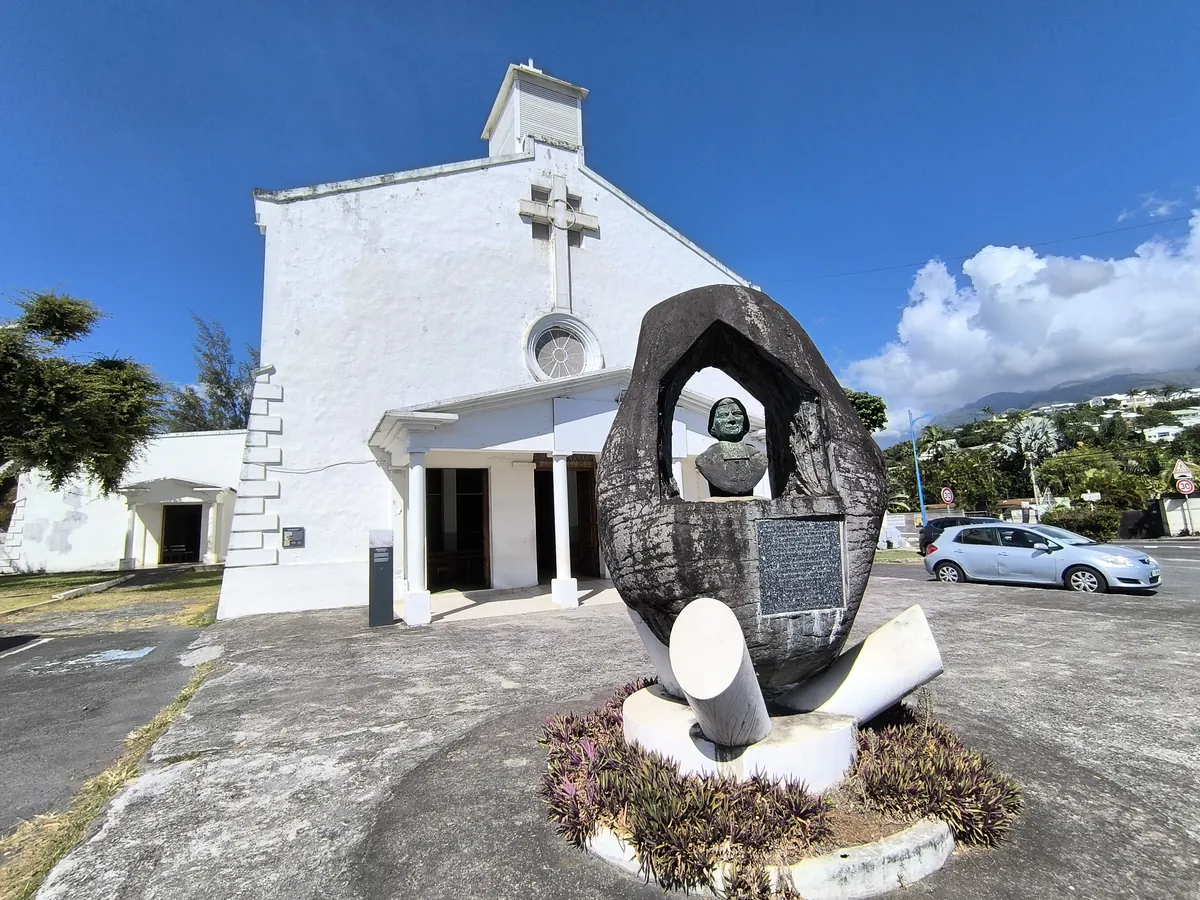 Monument en pierre sculptée sur le parvis bitumé devant l'église Saint-François-Xavier avec végétation et ciel bleu.