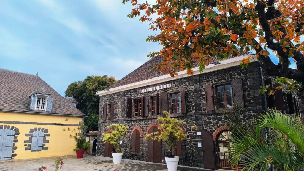 Façade en pierre de la mairie de Saint-Leu cadrée sous le feuillage d'un arbre aux feuilles rouges.