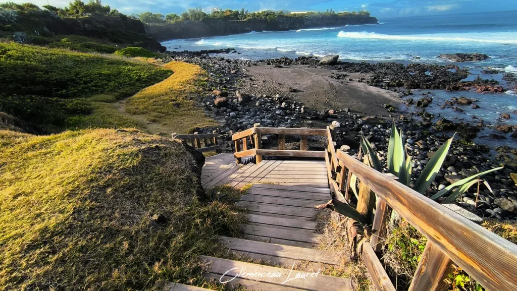 Vue en plongée d'un escalier en bois longeant une pente verdoyante et menant à une plage de sable noir et galets au bord du littoral.
