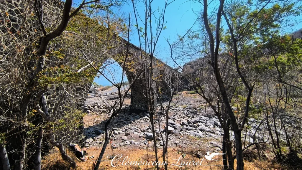 Vue à travers les arbres du pont ferroviaire en pierre de basalte de la ravine Colimaçon au-dessus d'un lit de rivière asséché.