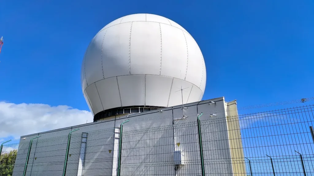 Gros plan sur le radôme sphérique blanc monté sur le bâtiment de la station météo sécurisée par un grillage.