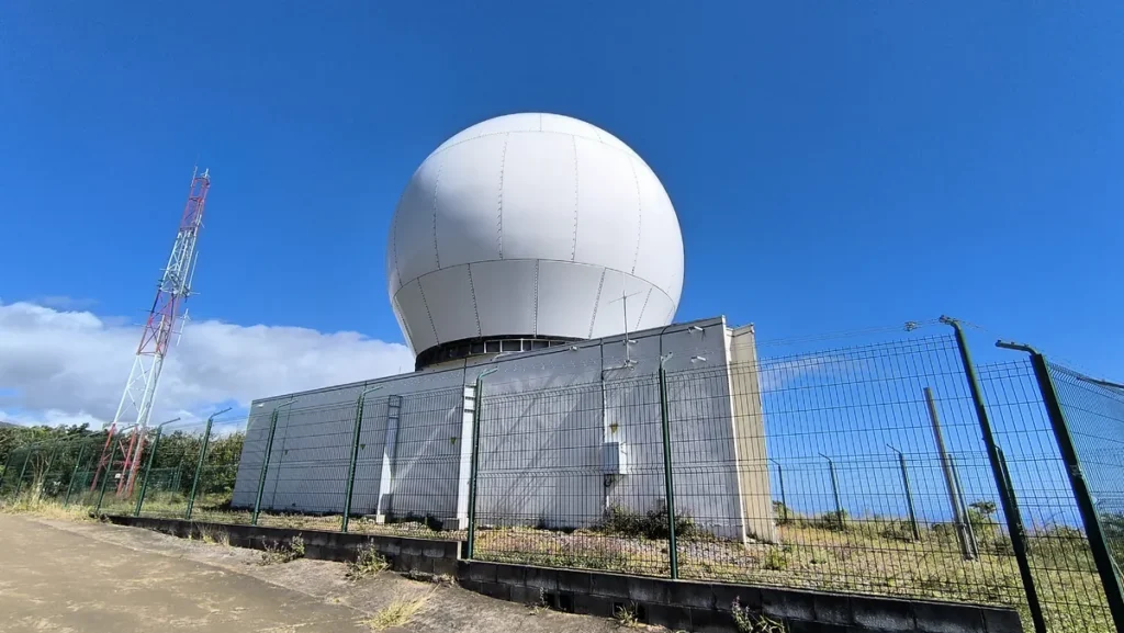 Vue en contre-plongée du bâtiment du radar météorologique avec le radôme blanc et un pylône de télécommunication métallique.