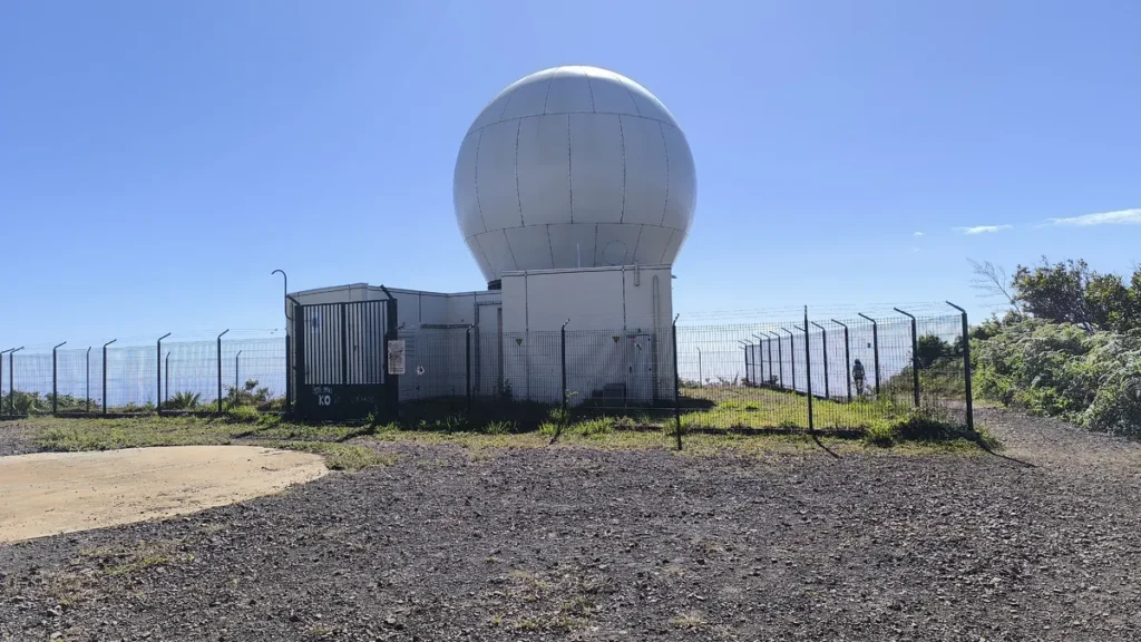 Vue latérale du complexe du radar météorologique avec portail noir et clôture métallique sur terrain gravillonné.