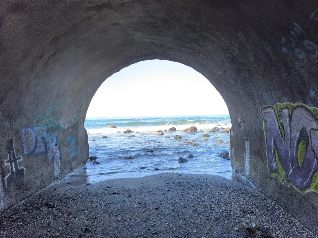 Cadrage centré sur les vagues et la plage de galets depuis l'ombre d'un tunnel tagué à la Ravine des Colimaçons, Saint-Leu.