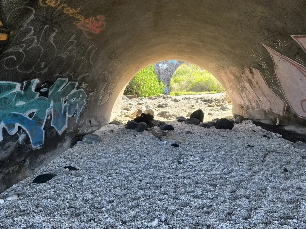 Sol en gravier et parois recouvertes de graffitis sous un tunnel ouvrant sur le viaduc de la Ravine des Colimaçons à Saint-Leu.