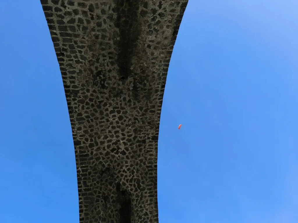 Vue en contre-plongée de l'arche en maçonnerie du viaduc de la Ravine des Colimaçons à Saint-Leu avec un parapente orange dans le ciel bleu.
