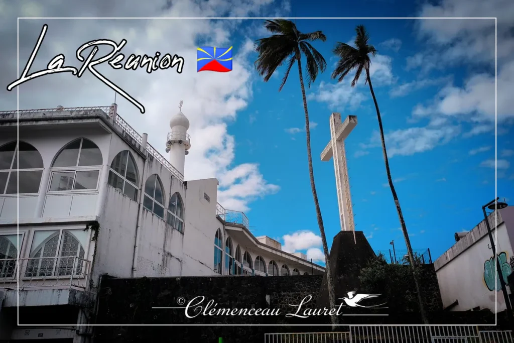 Vue en contre-plongée d'une mosquée blanche et d'une grande croix chrétienne en béton sous deux cocotiers et un ciel bleu nuageux à Sainte-Clotilde à La Réunion.