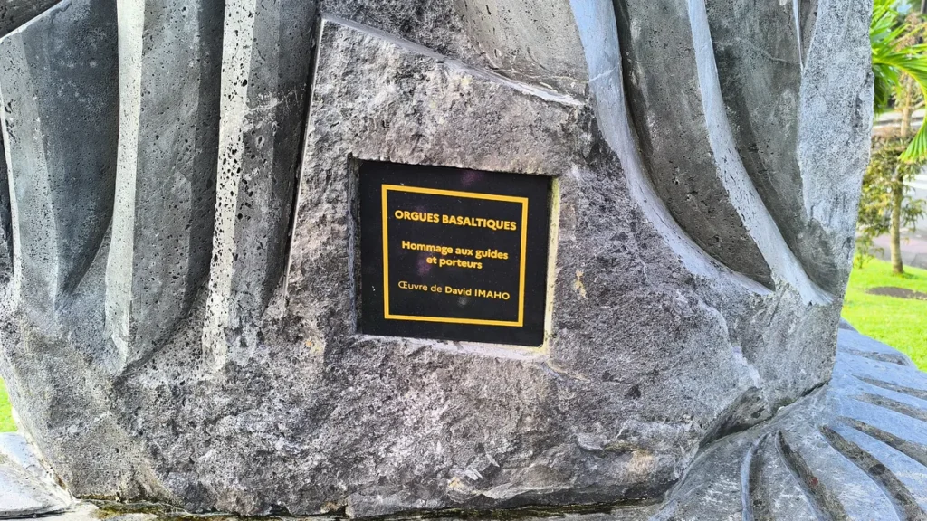 Plaque en métal noir gravée intégrée dans la roche indiquant Hommage aux guides et porteurs par David Imaho.