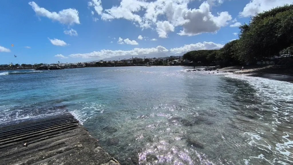 Plan d'eau turquoise scintillant sous le soleil, bordé par la cale en béton et la côte boisée de Saint-Pierre.