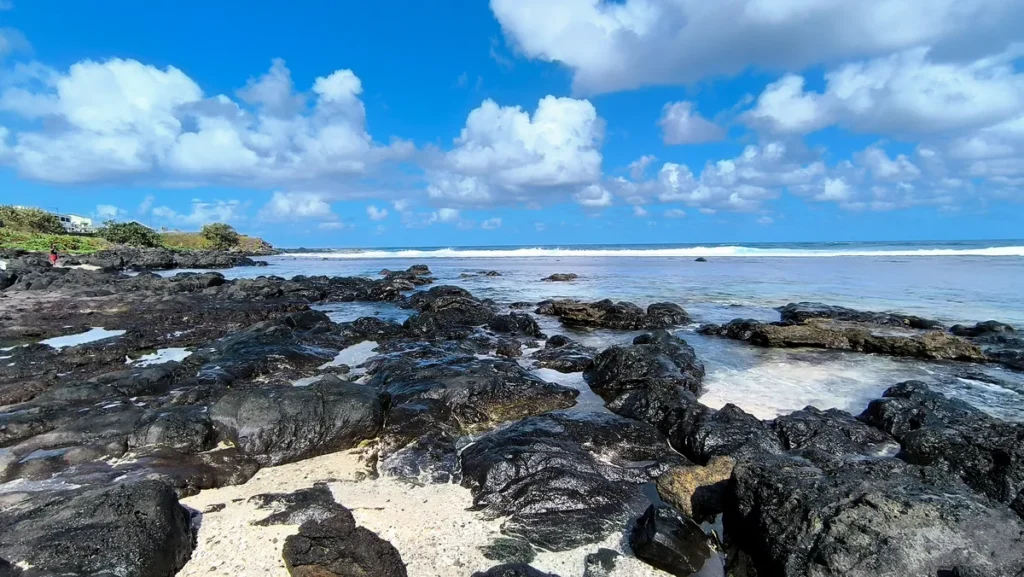 Étendue de roches volcaniques sombres formant des cuvettes d'eau de mer le long du littoral sous un ciel parsemé de cumulus.