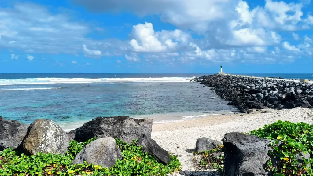 Jetée en enrochement menant au phare vert de Saint-Pierre vue depuis une plage bordée de végétation tropicale.