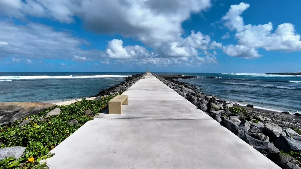 Longue promenade bétonnée rectiligne de la digue du port avec bancs, menant vers le phare entre deux étendues d'eau.
