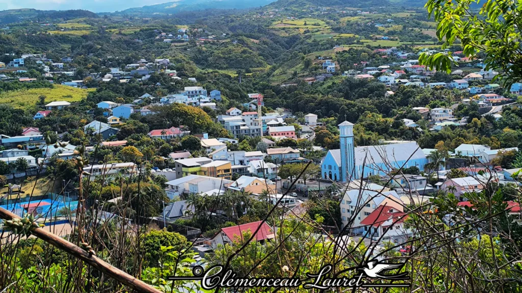 Plan rapproché plongeant sur l'église bleu ciel de Petite-Île et les toitures du bourg, encadré par des branches végétales au premier plan sous un ciel lumineux à La Réunion.