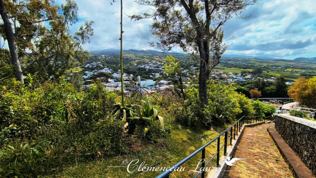 Vue en contre-plongée depuis un sentier pavé bordé d'une garde-corps métallique descendant vers la ville de Petite-Île avec une haute hampe d'agave au premier plan.