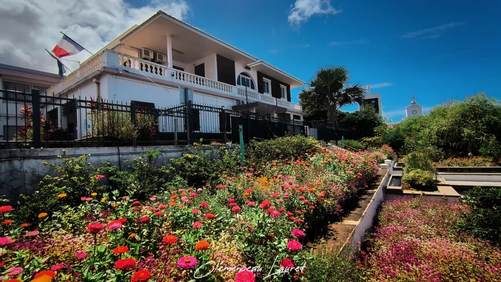 Vue en contre-plongée depuis le bas des jardins fleuris montrant la façade de la Mairie des Avirons surmontée du drapeau tricolore français, entourée de végétation tropicale, de palmiers et d'un tapis de fleurs sous un ciel bleu dégagé. Photo : Clémenceau Lauret.