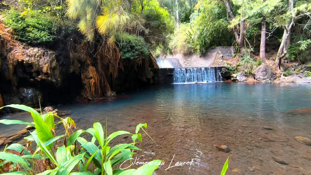 Bassin limpide aux reflets bleutés dominé par une cascade et une paroi rocheuse ferrugineuse orangée à Cilaos sous le soleil