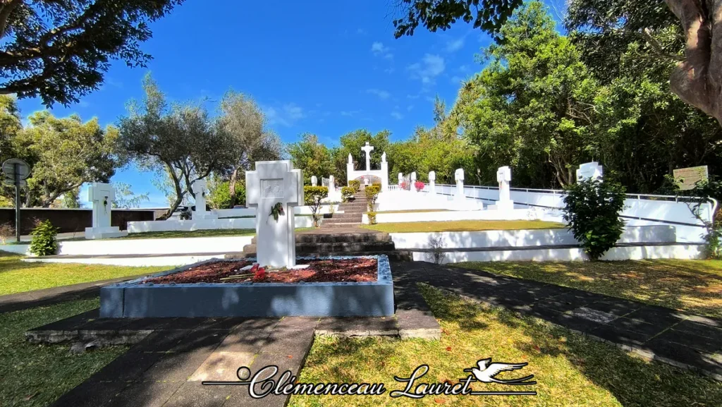 Chemin de croix et calvaire blanc monumental au site de Petite-Île à La Réunion. Vue en contre-plongée depuis le bas de l'allée montante. Au premier plan, une croix blanche sculptée est érigée sur un socle en pierre foncée garni de paillage rouge. Des escaliers en pierre et une rampe d'accès blanche mènent aux différentes stations du chemin de croix disposées en paliers successifs jusqu'à la grande croix centrale au sommet, entourée d'une végétation dense sous un ciel bleu limpide. - Photo : Clémenceau Lauret (Licence CC BY-SA 4.0)